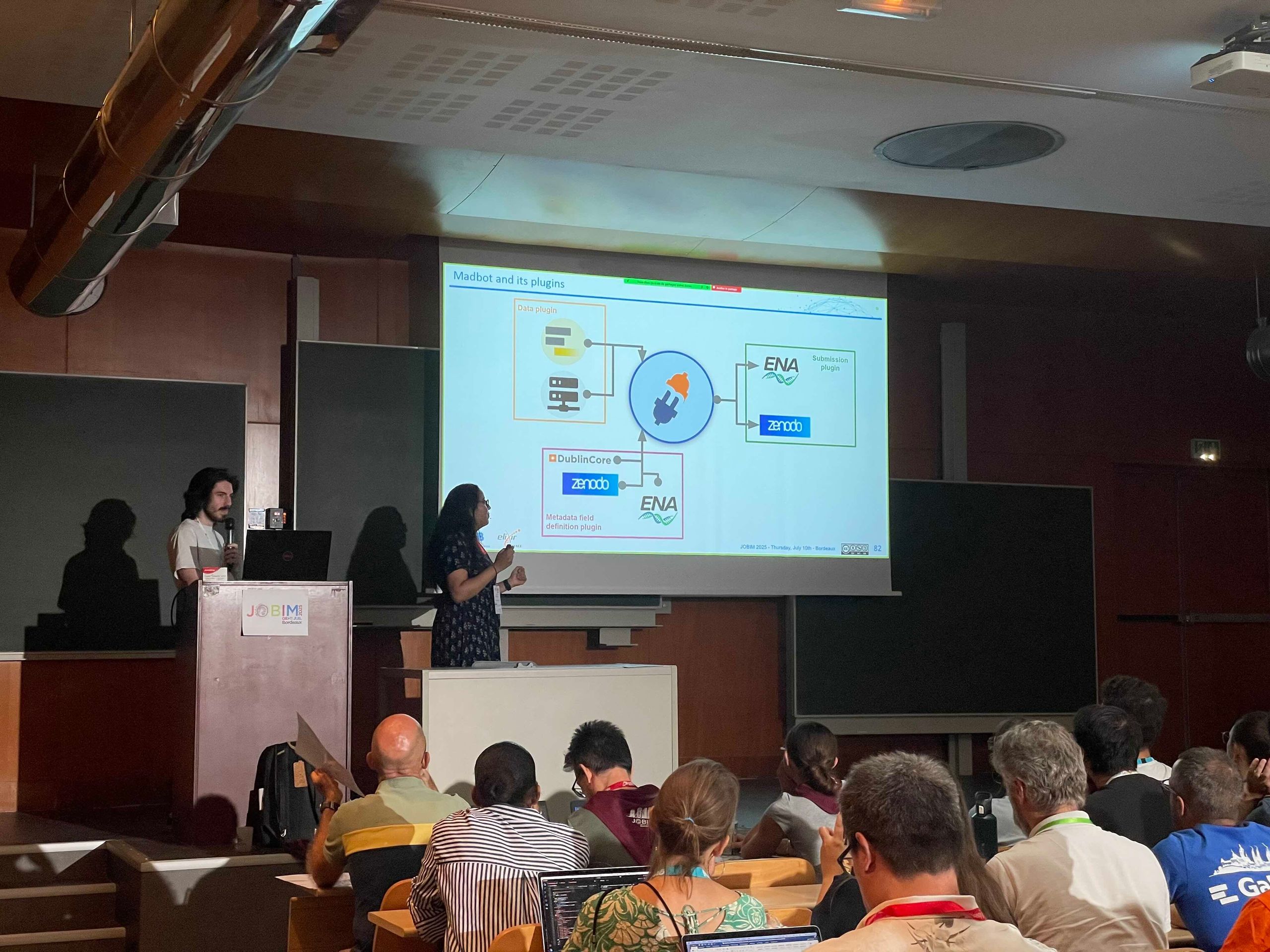 The image depicts a scene from a conference presentation. Two presenters are standing at the front of a lecture hall, actively engaged in delivering their talk. The presenter on the left is standing behind a podium with a laptop, while the presenter on the right is gesturing towards a projected slide on a large screen behind them. The slide is titled 'Madbot and its plugins' and features a diagram illustrating the interaction between different components such as a data plugin, DublinCore, Zenodo, and ENA (European Nucleotide Archive). The audience, seated and facing the presenters, appears attentive and engaged, with some individuals using laptops. The setting suggests a professional and academic environment focused on scientific or technical discussions.